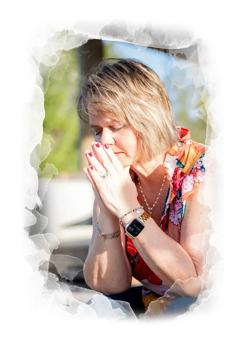 Kim Walker with short blonde hair sits with her hands clasped near her face, eyes closed, looking thoughtful or distressed. She wears a colorful top, a smartwatch, and has red nail polish. The background is blurred.