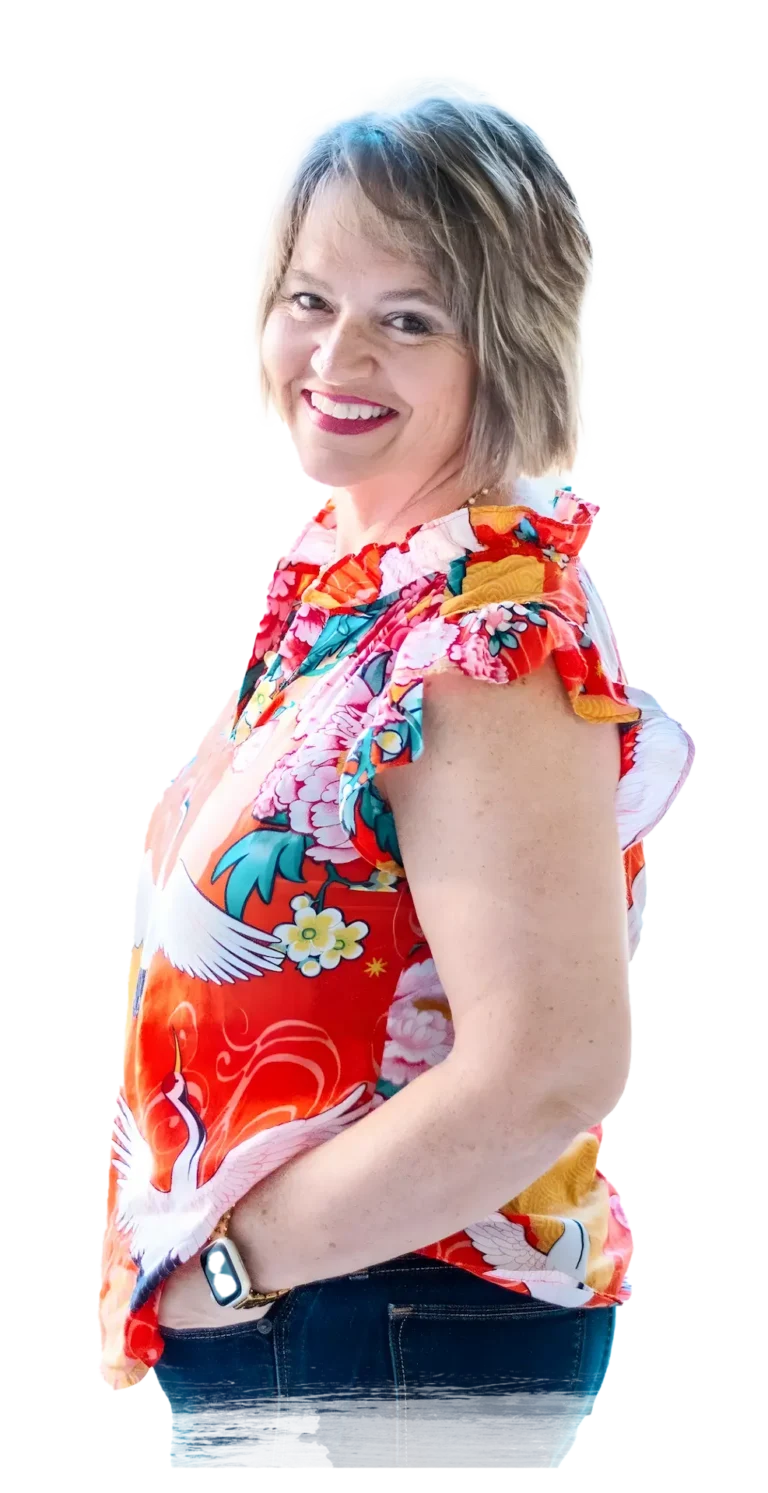 Image of Kim Walker with short, light brown hair stands sideways, wearing a colorful floral blouse with white cranes and blue jeans, her hand in her pocket.