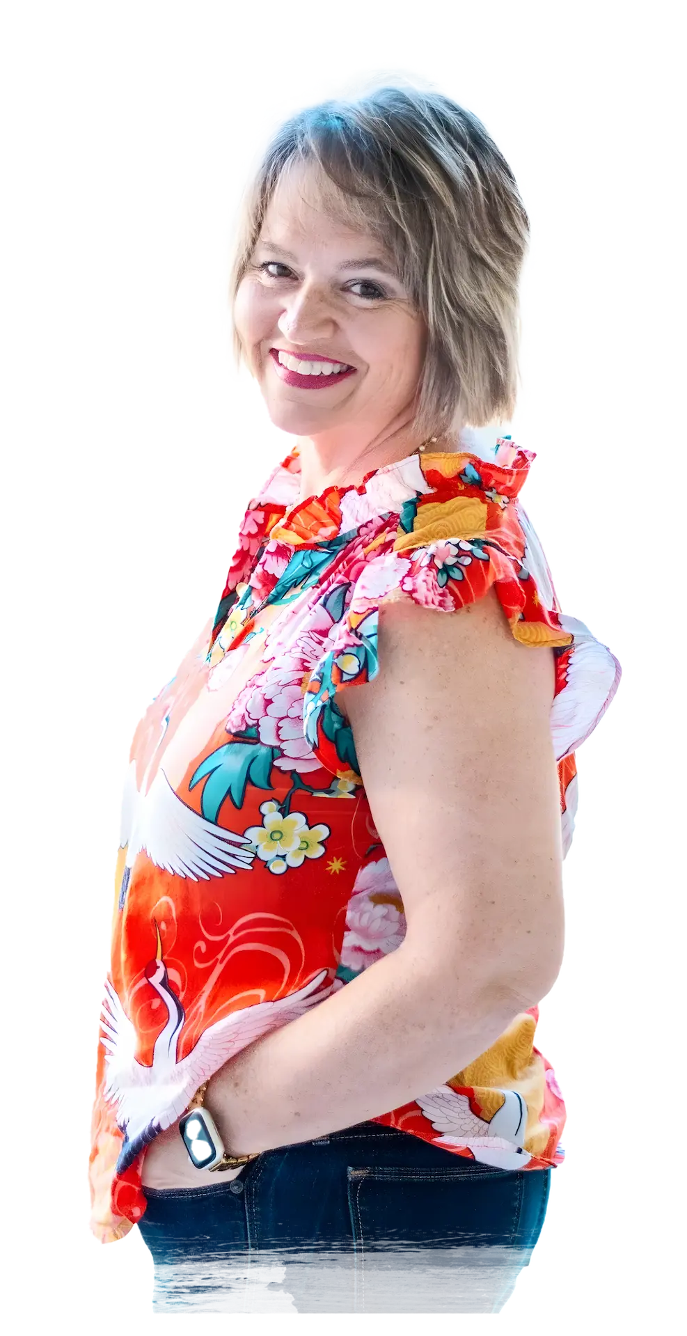 Image of Kim Walker with short, light brown hair stands sideways, wearing a colorful floral blouse with white cranes and blue jeans, her hand in her pocket.