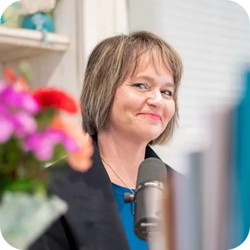 Kim Walker with short light brown hair and a blue top smiles slightly while sitting behind a microphone. Colorful, out-of-focus flowers appear in the foreground. Shelves with decor are visible in the background.