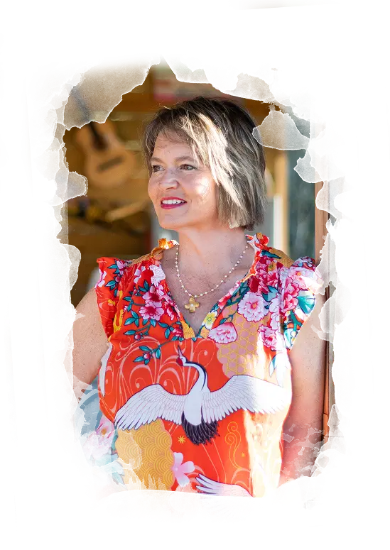 Podcast, Image of Kim Walker with short brown hair stands smiling near a doorway, wearing a bright orange floral blouse with white cranes. A guitar is visible in the blurred background.