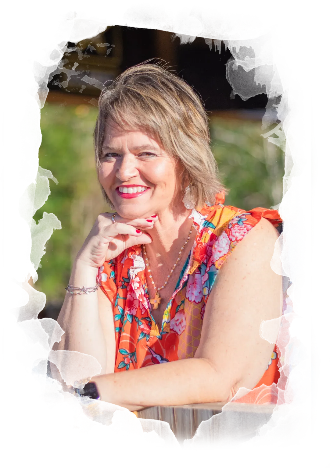 About, Kim Walker smiling with short light brown hair, wearing a bright floral sleeveless top, red lipstick, and an Apple Watch, poses outdoors with her chin resting on her hand.