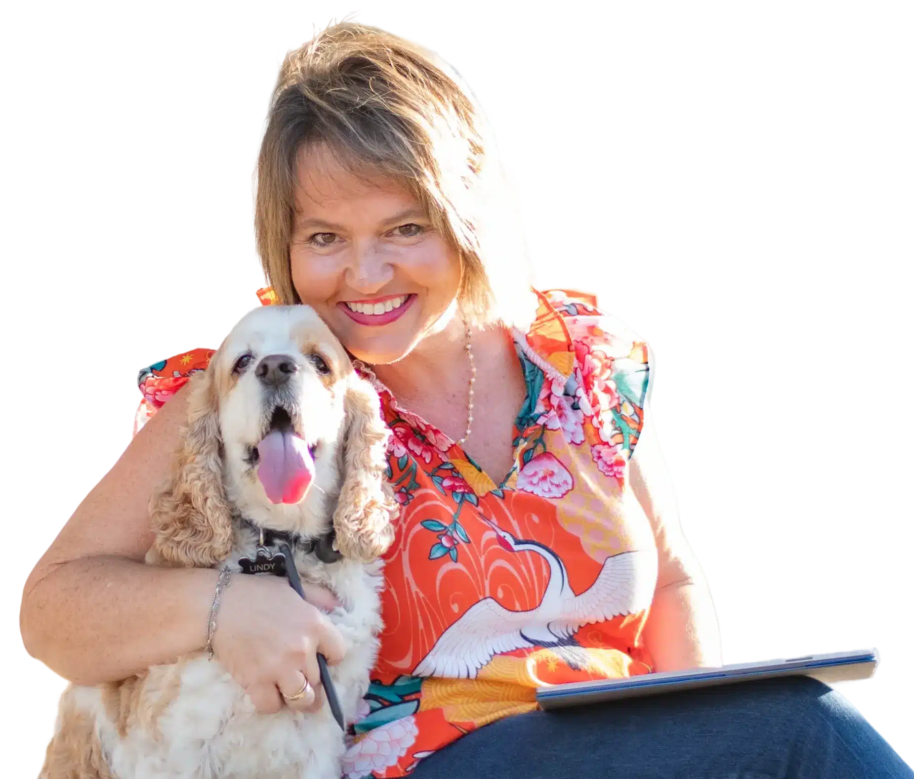 An image of Kim Walker in a colorful shirt sits with a happy, tan and white dog. The dog is panting and looking at the camera. The woman is holding the dogs paw and has a tablet on her lap.