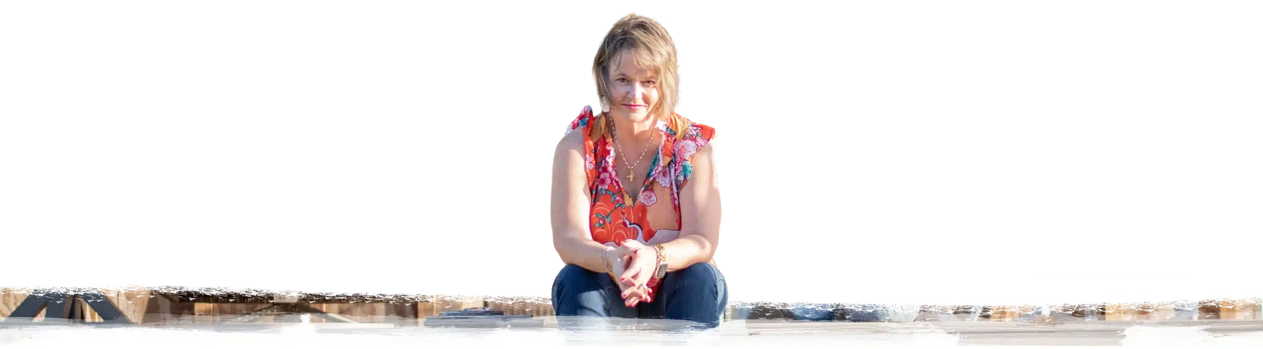 Kim Walker with light hair wearing a colorful sleeveless top and jeans sits outdoors on a low surface, hands clasped, looking toward the camera, with a dark background behind them.