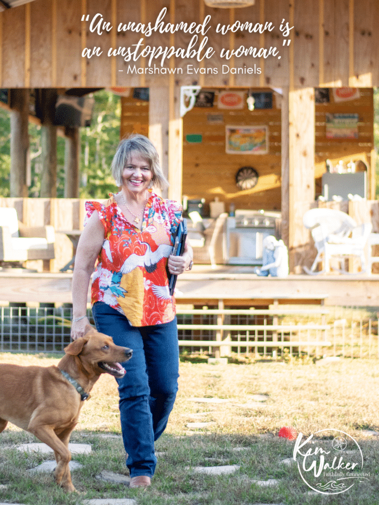 Kim Walker in a colorful blouse holds a gun and walks outside with a golden retriever. A rustic wooden cabin is in the background. A quote above her reads, “An unashamed woman is an unstoppable woman.”.
