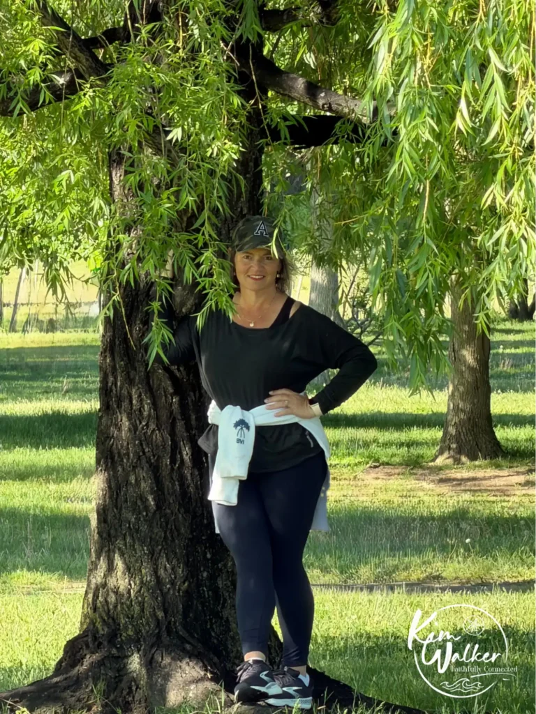 Kim in black overall with a cap and a white jacket wrapped around her waist under of a weeping willow tree.