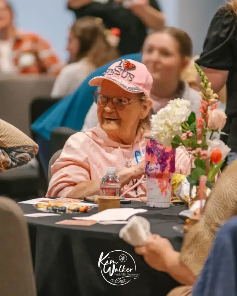 An elderly woman in a pink hoodie and cap sits at a table with flowers, snacks, and a water bottle, smiling at someone off camera. Other people are visible in the background.